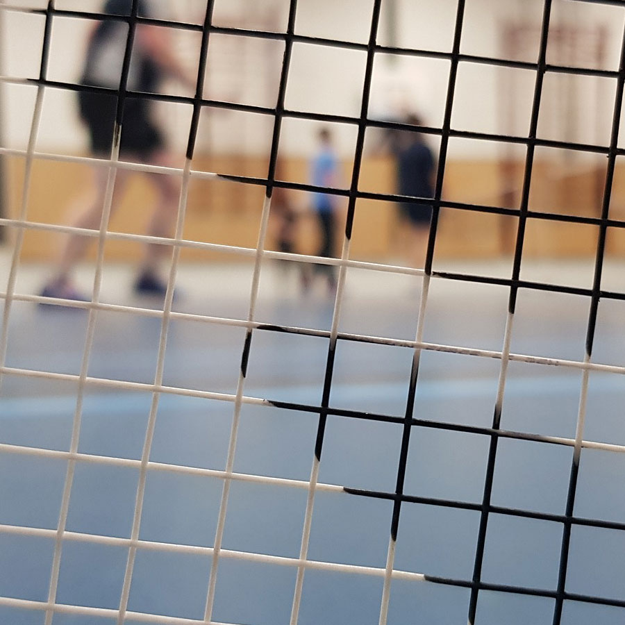Close view through a badminton net in the Drill Hall, with players out of focus on the blue court.