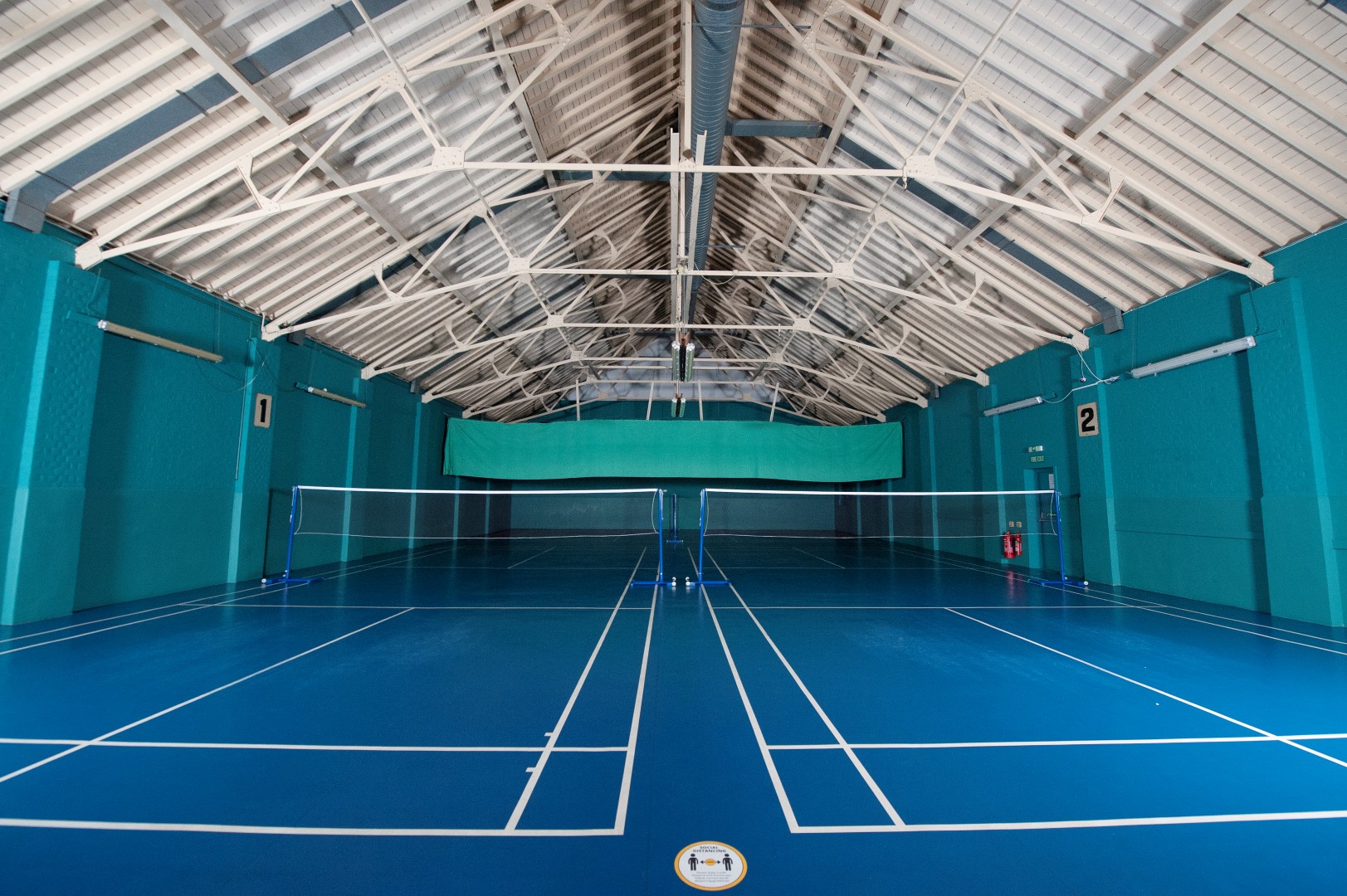 The main hall at Enfield Drill Hall: blue badminton courts, white lines, teal walls and a high roof of white metal trusses.