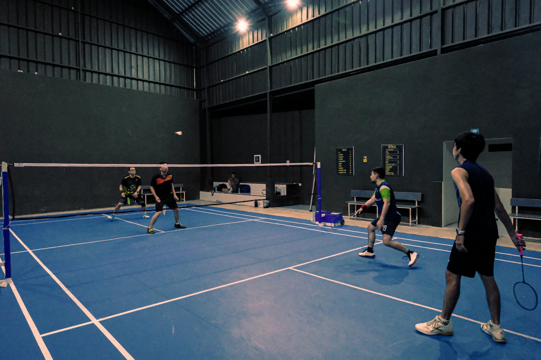 Four adults playing a doubles rally on an indoor badminton court.